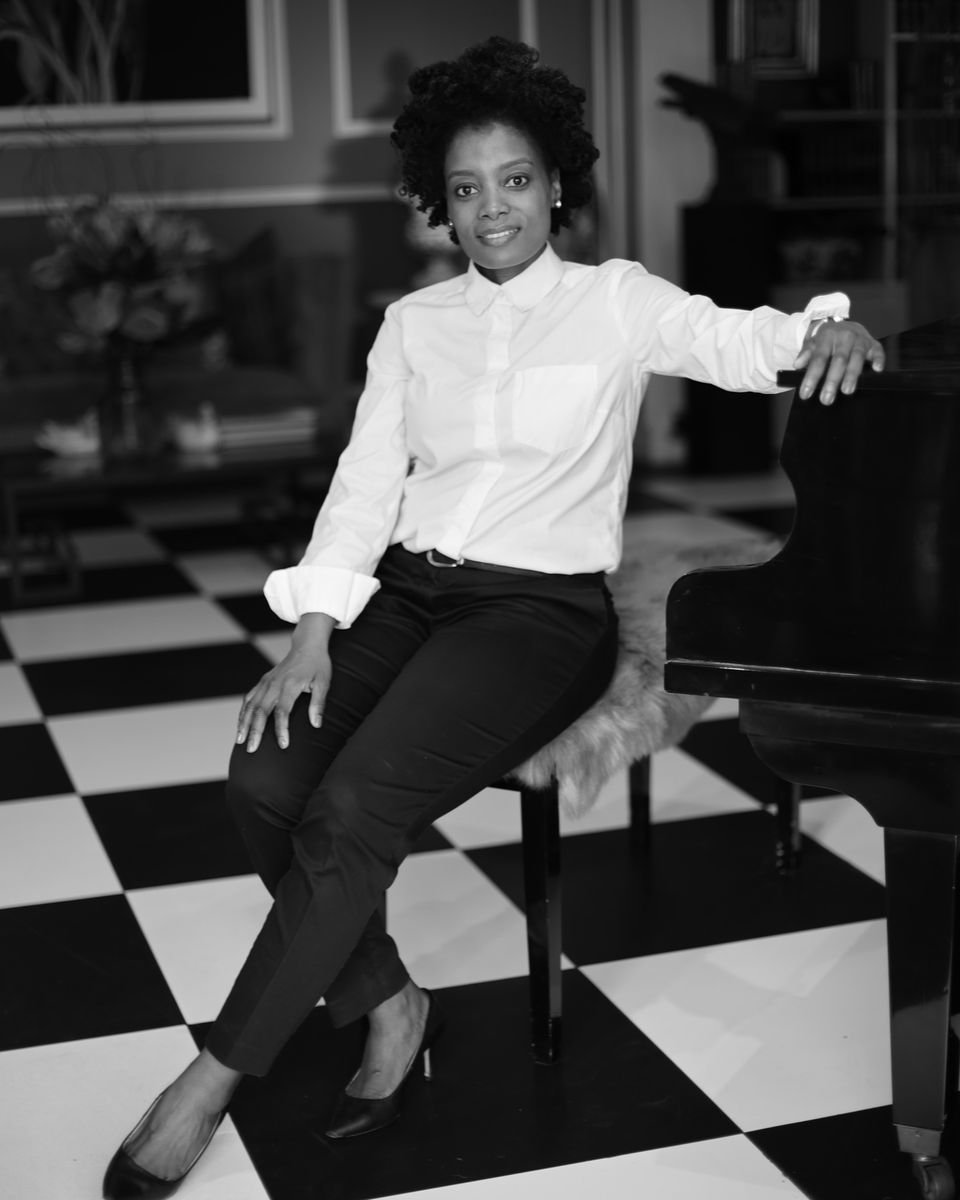 Maud Nale seated beside a grand piano on a chequered floor, in black and white